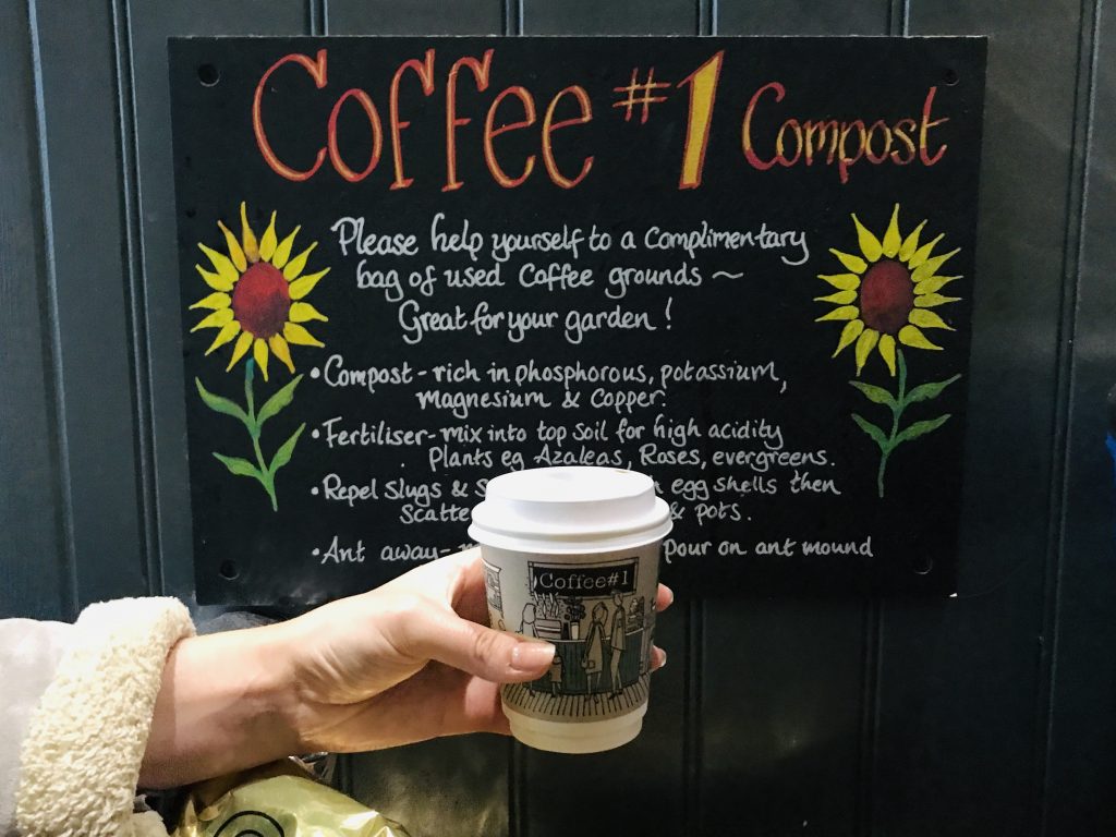 Coffee cup being held up beside a Coffee #1 poster about compost