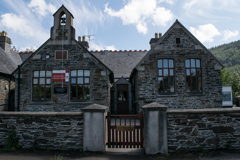 An old stone building faces towards the viewer, surrounded by a waist-high stone gate. A red sign on the left reads: "Bunscoill Ghaelgagh"