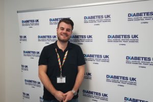 Josh James who works for the Diabetes UK stands in front of a a backdrop showing the charity's logo. 